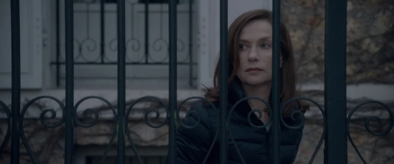 Elle (2016) movie still: medium — A woman stands behind a black wrought iron gate with swirling decorative details. The background shows a portion…