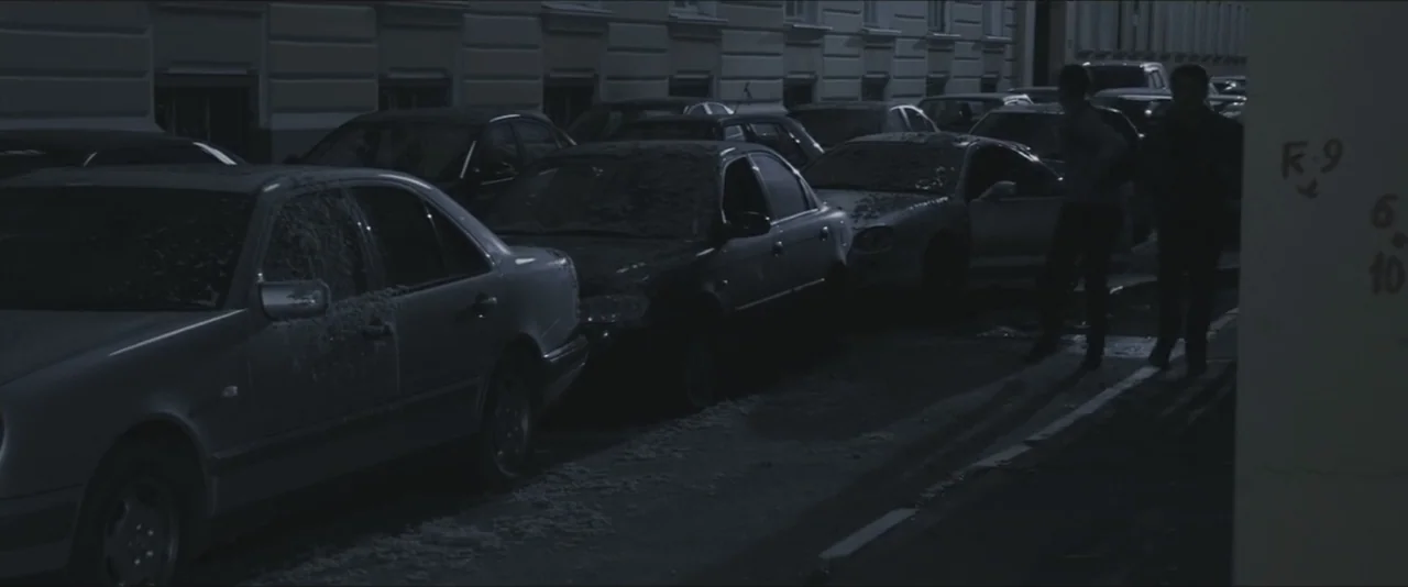 Darkest Hour (2017) movie still: wide — A dark city street at night is lined with multiple cars parked in rows. Two men stand together near the…