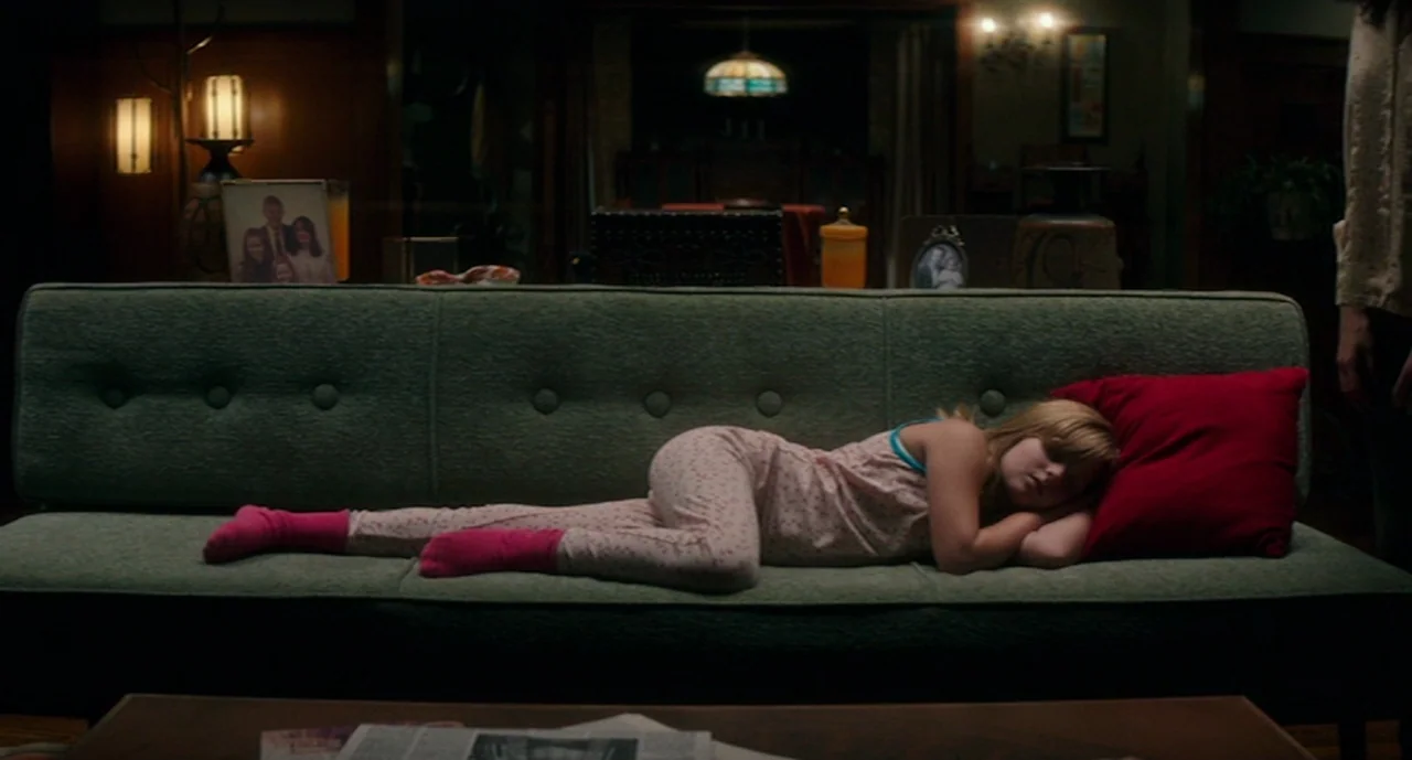 Ouija: Origin of Evil (2016) movie still: medium — A dimly lit room features a mid-century style green sofa against a wooden wall. A young girl in…