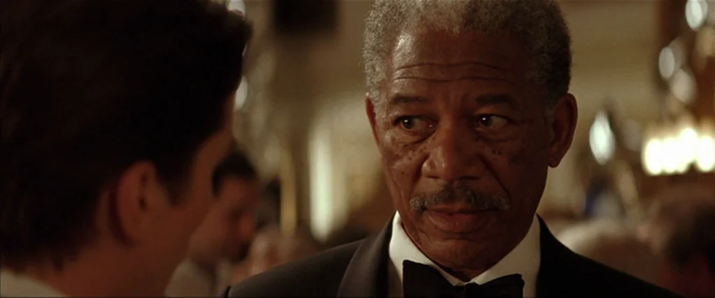 Batman Begins (2005) movie still: closeup — A well-lit, busy interior space features warm tones and blurred light fixtures in back. An older man in…