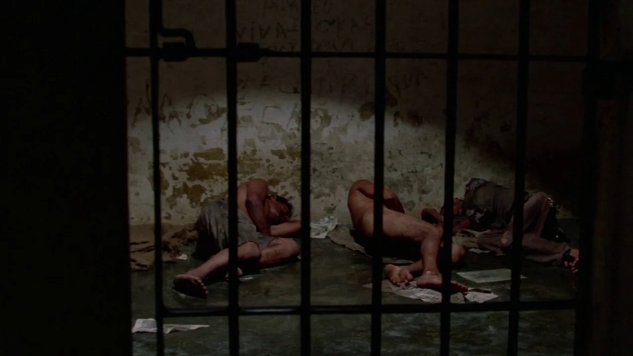 Sorcerer (1977) movie still: wide — A cramped prison cell with grimy walls featuring scratched graffiti and concrete floors. Three men lie on the…
