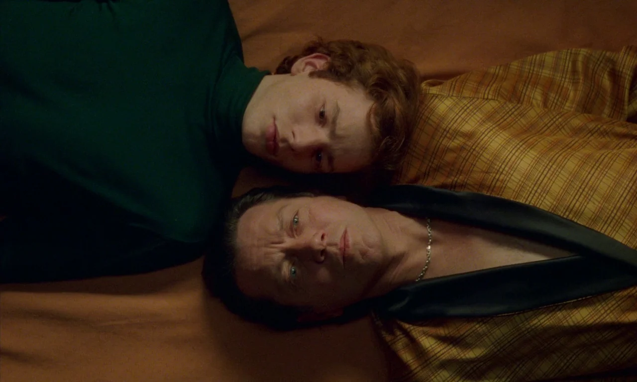 Water Drops on Burning Rocks (2000) movie still: closeup — Two men lie on their backs on an orange surface. They gaze upward while positioned next…