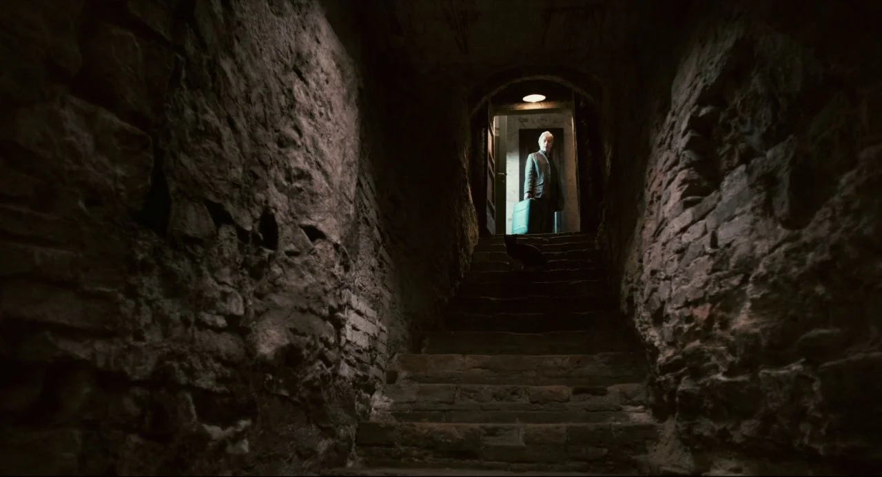 Certified Copy (2010) movie still: wide — A dark, narrow stone staircase leads up to a brightly lit doorway at night. An elderly man in a suit…
