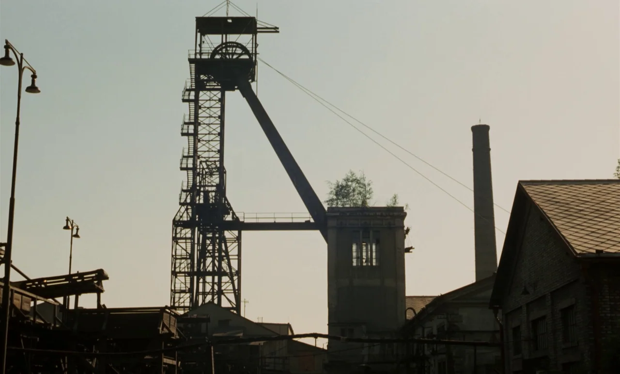 Larks On A String (1969) movie still: establishing — A massive metal mine headframe rises prominently above a collection of low, dark industrial…
