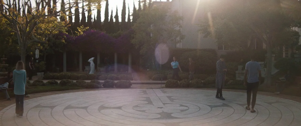 Honey Boy (2019) movie still: wide — A stone paved outdoor labyrinth surrounded by hedges and tall cypress trees at dusk. Several people walk across…