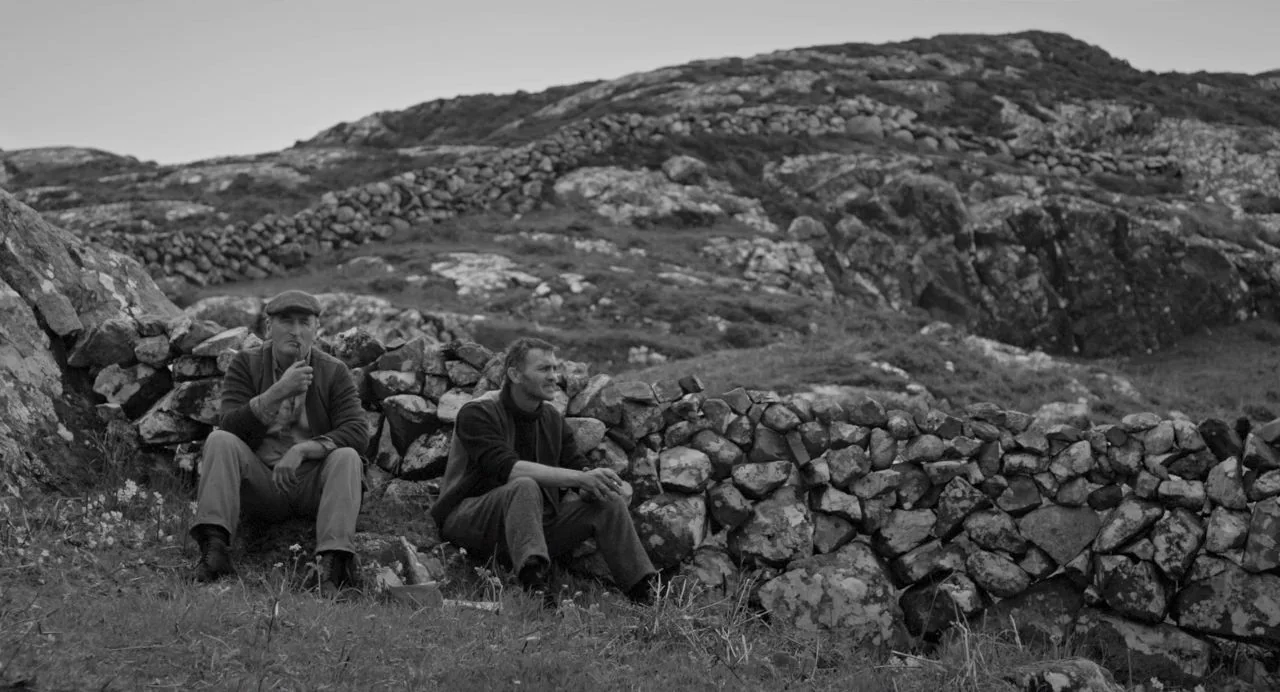 Song of Granite (2017) movie still: wide — A rocky, windswept Irish landscape features a dry stone wall running through the frame. Two men sit on…