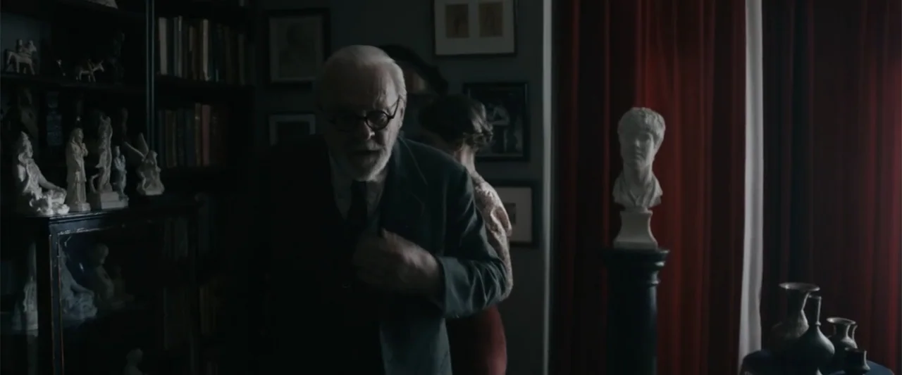Freud's Last Session (2023) movie still: medium — A dimly lit room filled with bookshelves, white statuettes, and dark red curtains. An elderly man…