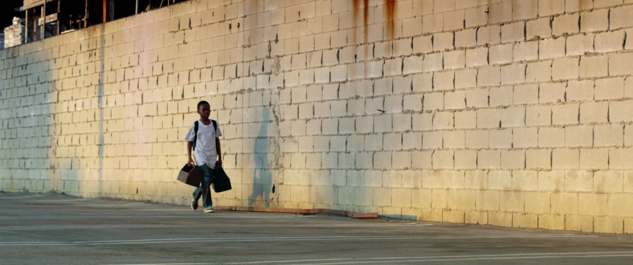 Ambulance (2022) movie still: wide — A long concrete block wall borders a paved lot at sunset. A young boy carries two boxes as he walks parallel to…