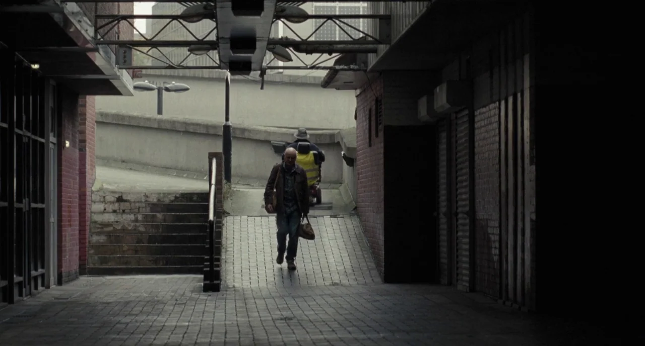 I, Daniel Blake (2016) movie still: wide — A concrete pedestrian ramp leads upward between brick walls under an overhead steel structure. A man…