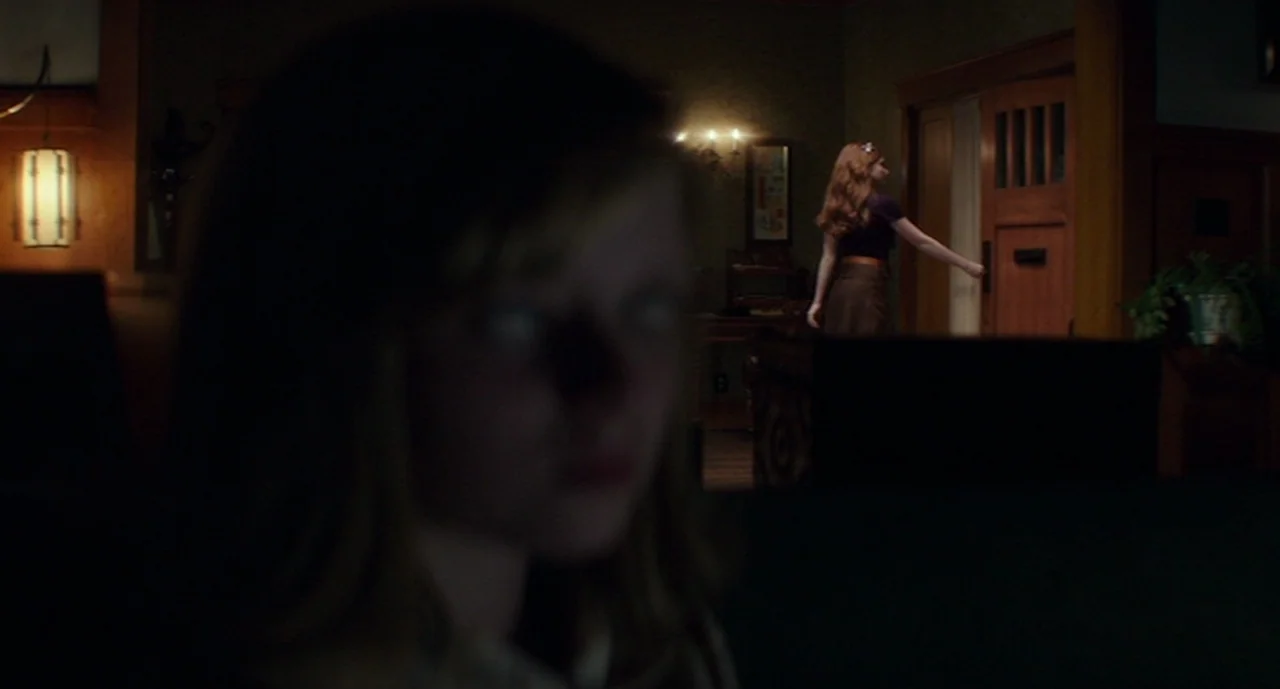 Ouija: Origin of Evil (2016) movie still: medium — A dimly lit room with dark wood trim and 1960s style decor. A woman in the background reaches…