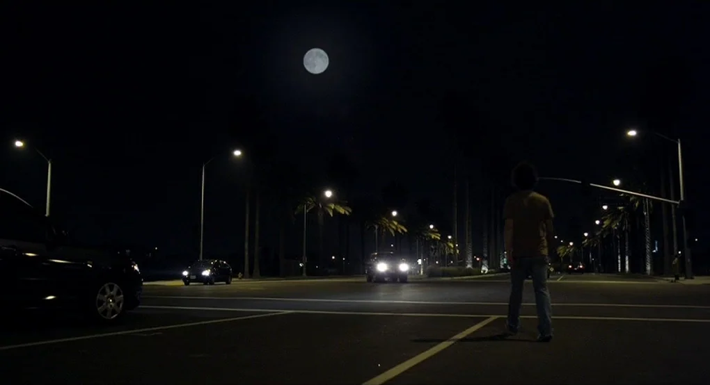 The Future (2011) movie still: wide — A dark, paved road intersection at night features palm trees and streetlights under a full moon. A lone man in…