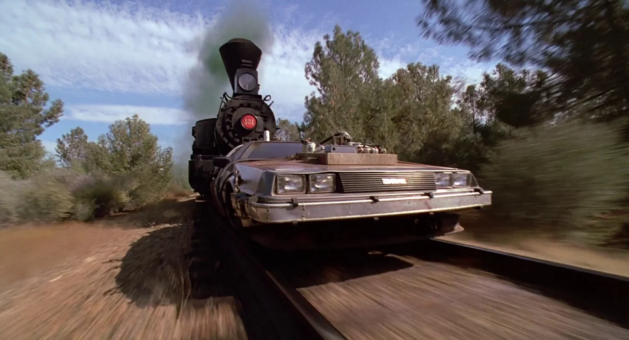 Back To The Future Part III (1990) movie still: wide — A steam train pushes a sports car mounted on custom wheels along wooden tracks. Pine trees…
