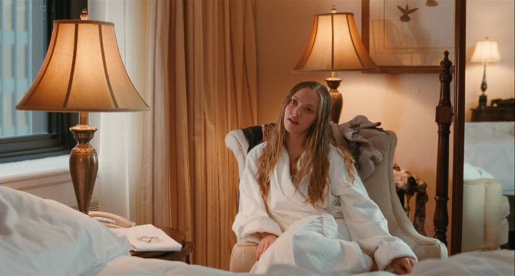 Chloe (2009) movie still: medium — A hotel room with beige walls and curtains illuminated by warm table lamps. A young woman with long hair sits in…