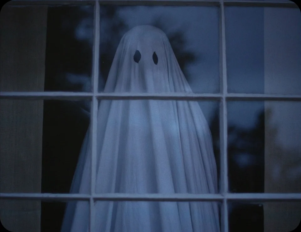 A Ghost Story (2017) movie still: medium — A person covered by a white sheet stands behind a window with grid muntins. The figure gazes forward…