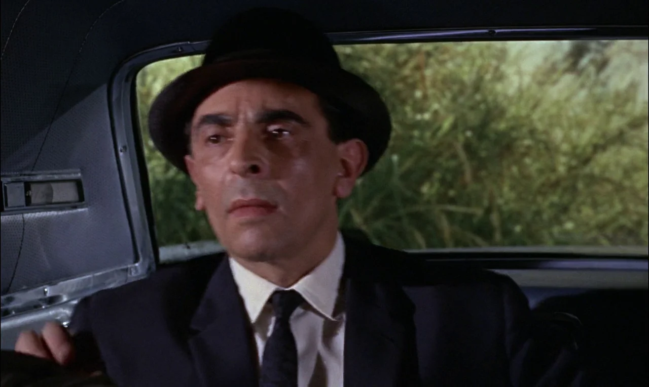 Goldfinger (1964) movie still: medium — The interior of a moving vehicle shows an adult man seated in the back. Outside the car window, green…