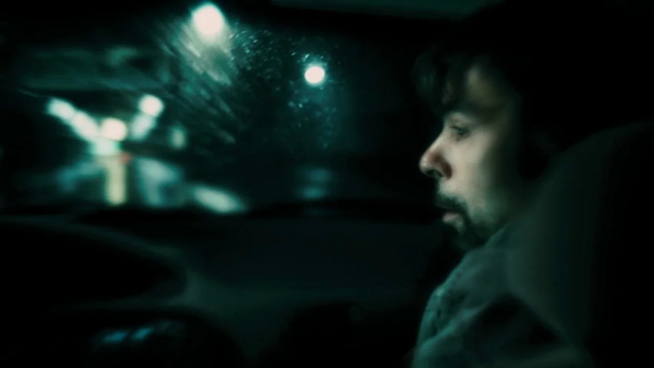 Saw VI movie still: closeup — The interior of a car at night during a rainstorm shows a blurred urban environment. A man sits in the passenger seat…