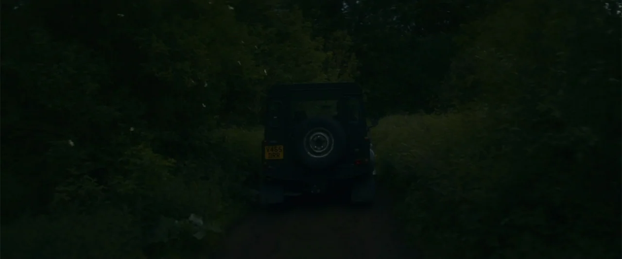 Black Bag (2025) movie still: medium — A dark off-road vehicle travels along a narrow dirt path flanked by dense foliage. The vehicle moves away…
