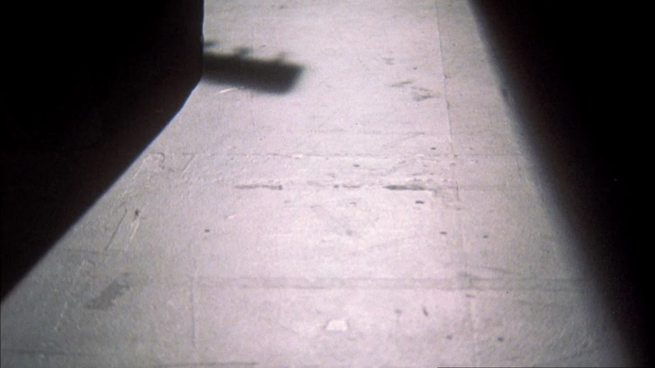 Stop Making Sense (1984) movie still: closeup — A light beam illuminates a portion of a textured, grey concrete floor surface. The sharp shadow of a…