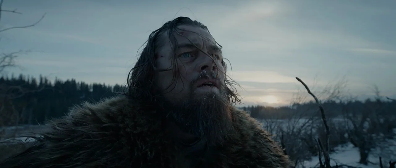 The Revenant (2015) movie still: closeup — A man stands in a desolate, snow-covered landscape during a winter sunset. The subject is a man with long…