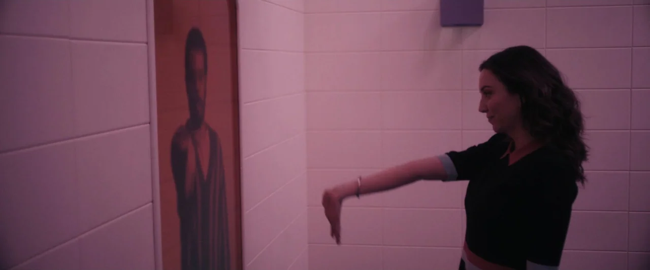 Sign Here (2017) movie still: medium — A tiled room features a glowing red vertical panel displaying a portrait of a man. A woman stands in the…