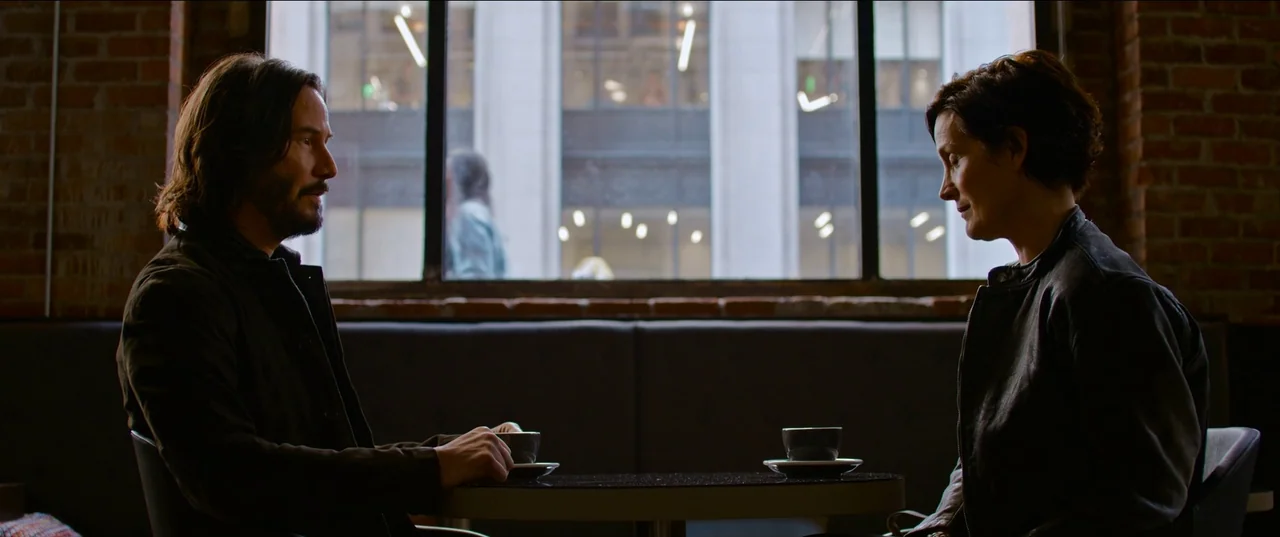The Matrix Resurrections (2021) movie still: medium — A dark coffee shop interior features brick walls and a large windowed rear view. A man and a…