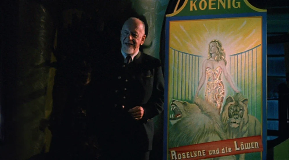 Roselyne and the Lions (1989) movie still: medium — An older man stands inside a dimly lit space next to a large poster. The poster depicts a woman…
