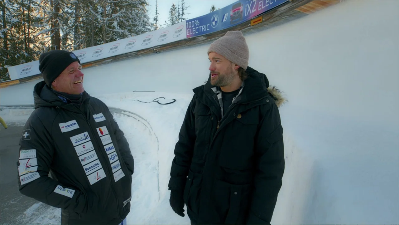 Jack in Time for Christmas (2024) movie still: medium — Two men stand within the curved ice walls of a professional bobsleigh track. The men face…