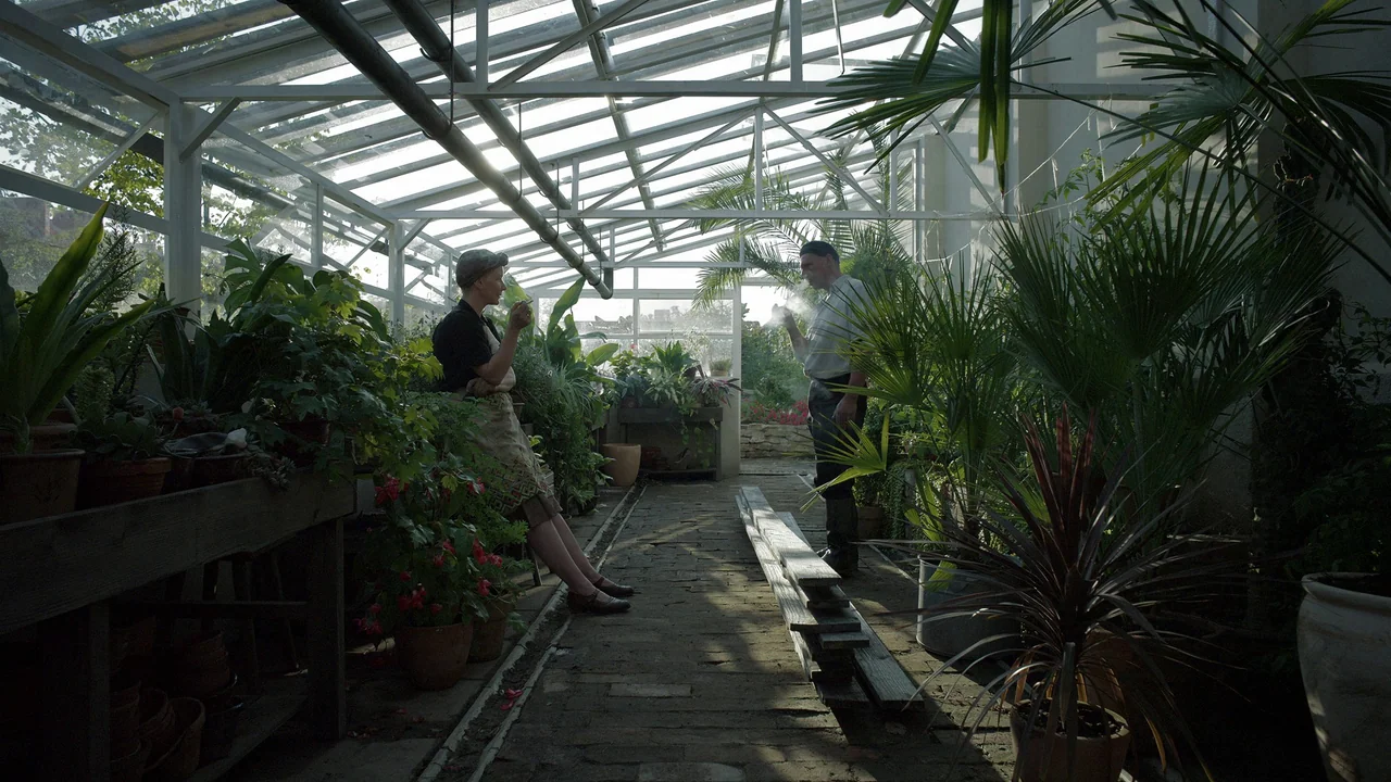 The Zone Of Interest (2023) movie still: wide — A glass-walled greenhouse contains numerous potted plants and a narrow brick walkway. A man and a…