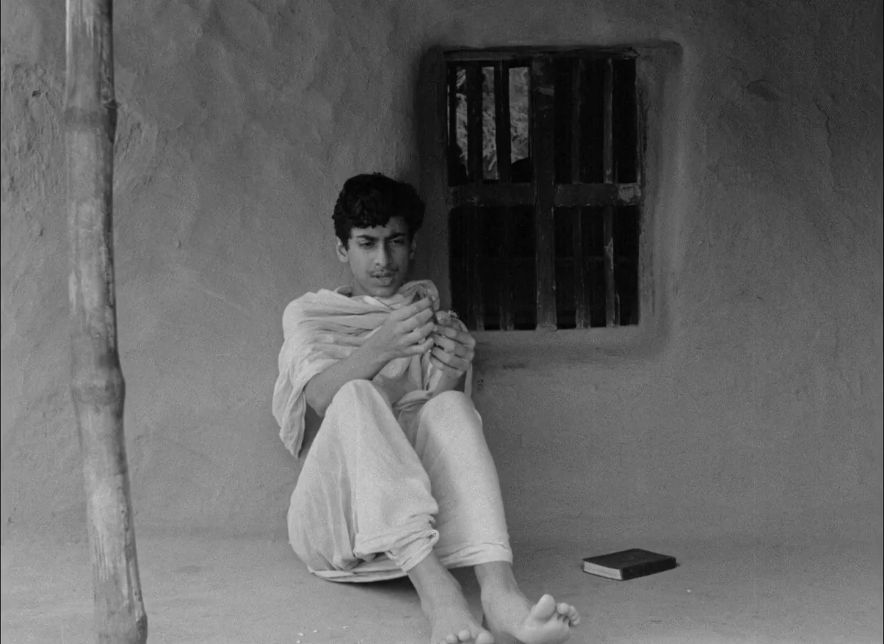 Aparajito (1956) movie still: medium — A rustic mud-walled exterior structure features a small window with dark vertical wooden bars. A young man…