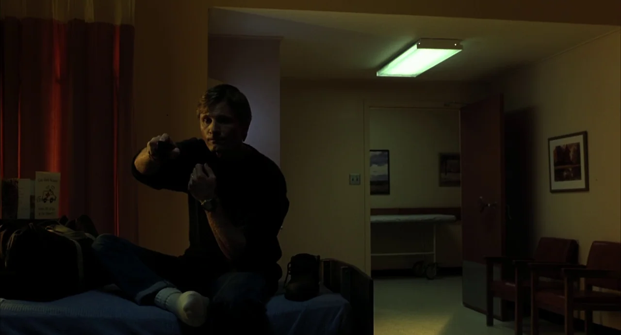 A History of Violence (2005) movie still: medium — A dimly lit room contains a bed, red curtains, and two leather chairs. A man sits on the edge of…