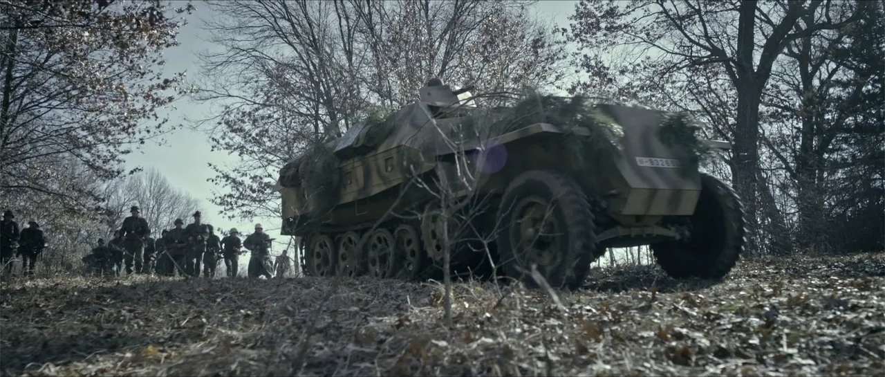 Battle of the Bulge (1965) movie still: wide — A German Sd.Kfz. 251 half-track drives through a sparse, leafless winter woodland. A small group of…