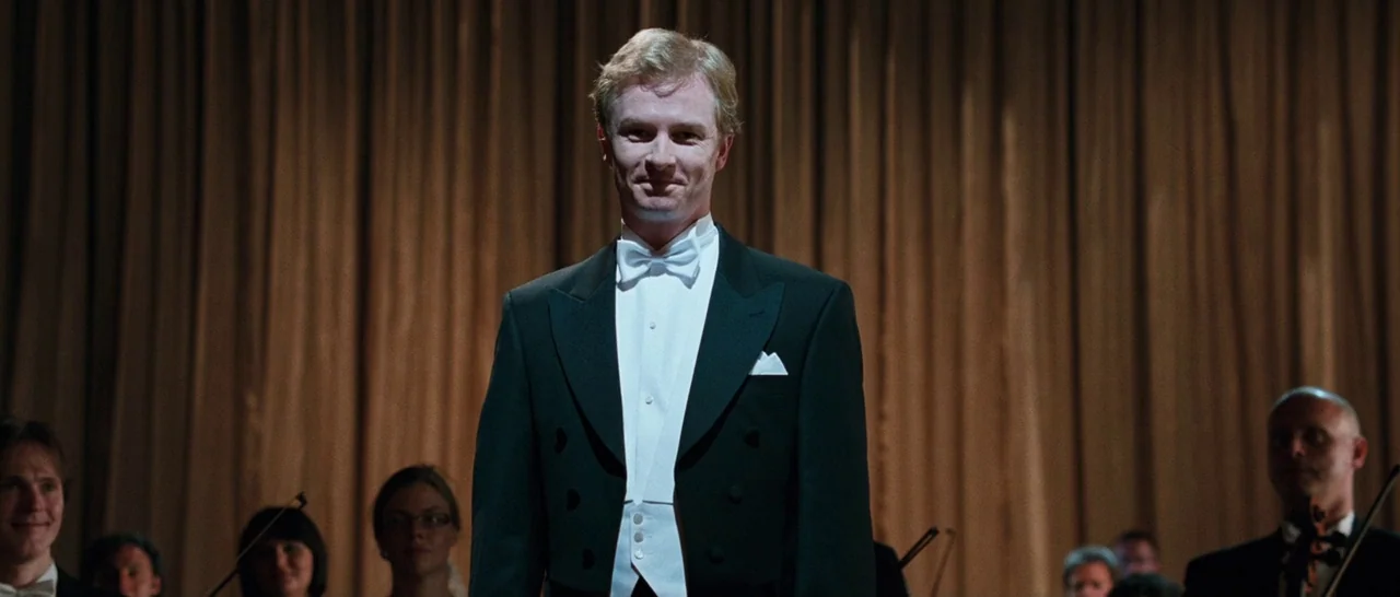 Sound of Noise (2010) movie still: medium — A man in a formal tuxedo stands in front of a golden curtain. Several musicians are partially visible in…