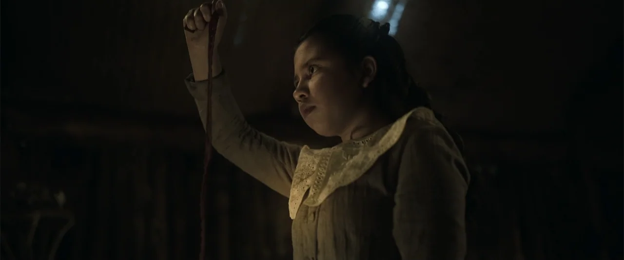 Evil Eye (2020) movie still: medium — A dark, dimly lit interior space with shadows obscuring the surrounding walls. A young girl in period clothing…