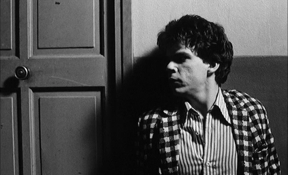 Boy Meets Girl (1984) movie still: medium — A hallway with a plain wall and a single paneled door frame. A man leans his shoulder against the wall…