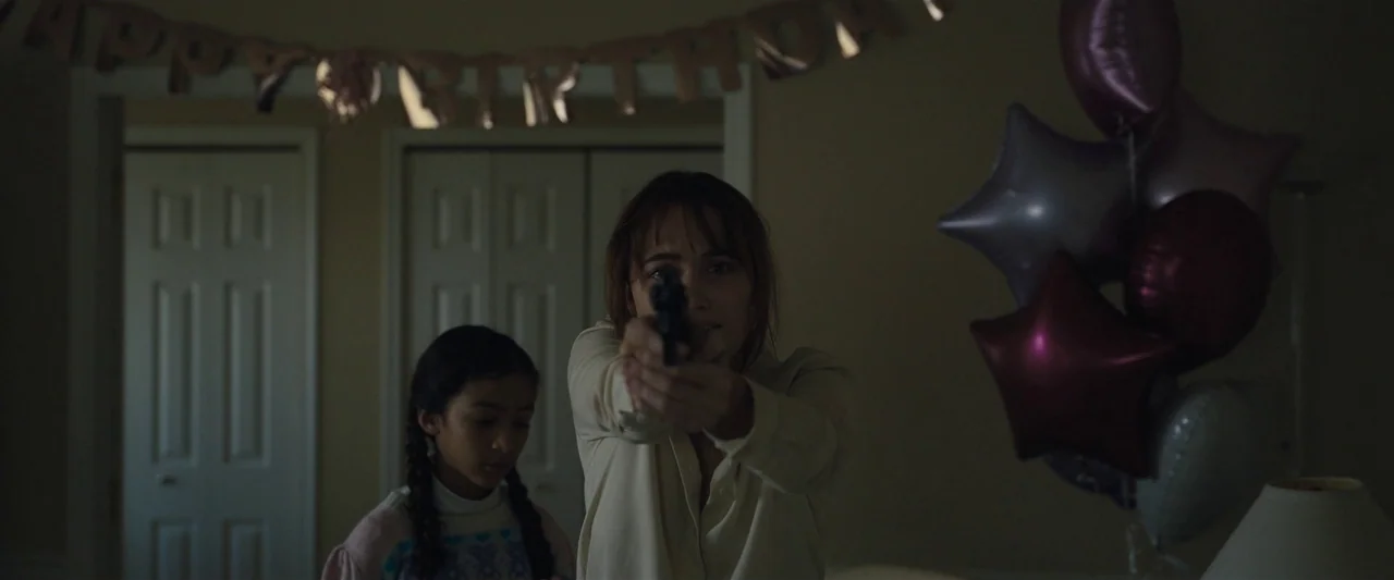 Longlegs (2024) movie still: medium — A dimly lit room displays a 'HAPPY BIRTHDAY' banner and clustered star-shaped balloons. A woman holds a…