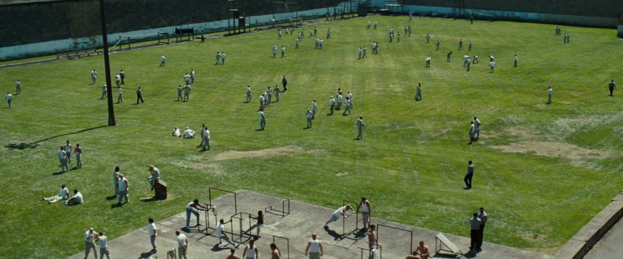 Out of the Furnace (2013) movie still: establishing — An expansive outdoor prison yard features a large grassy field and concrete exercise area.…