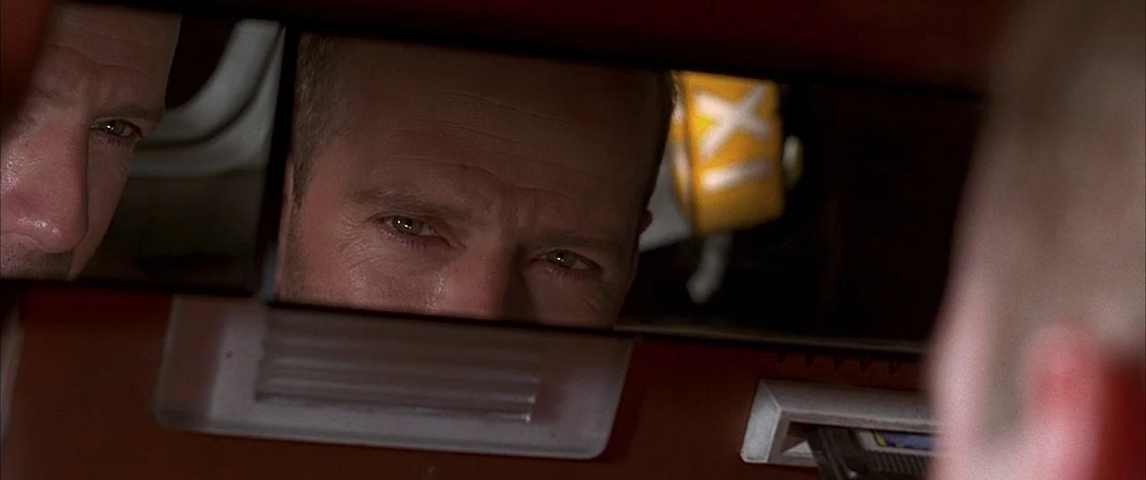 The Fifth Element (1997) movie still: closeup — The interior of a 1990s vehicle cabin at night with a plastic rearview mirror. A man is reflected in…