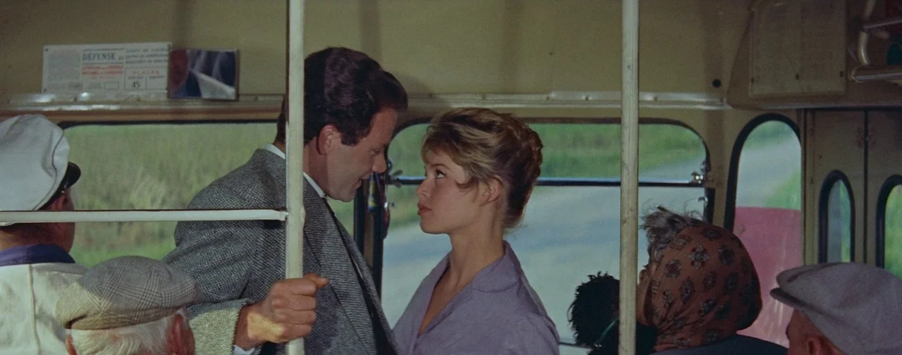 And God Created Woman (1956) movie still: medium — A crowded public bus interior features vertical metal poles and blurred rural scenery outside. A…
