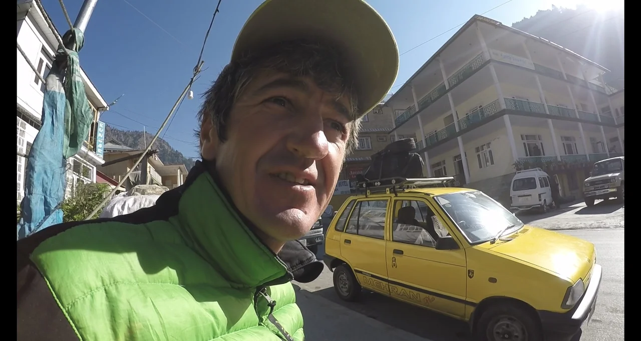 Blutch (2018) movie still: closeup — A sunny village street with a yellow car parked in the background. A man looks toward the side while wearing a…