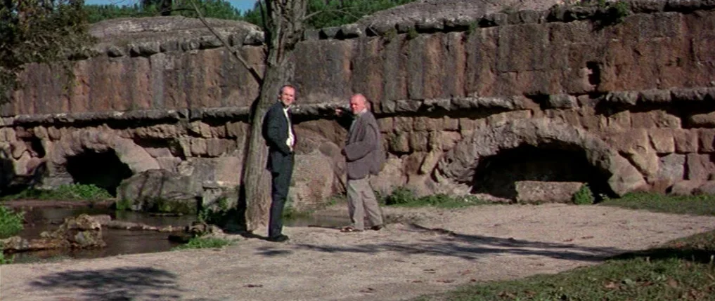 Titus (1999) movie still: fullbody — Two men stand before a long, ancient stone wall with arched openings. A narrow stream flows in the foreground…