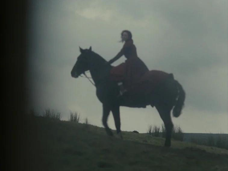 Wuthering Heights movie still: wide — An overcast moorland landscape features sparse grass and a darkened, cloudy sky. A woman in a red dress rides…
