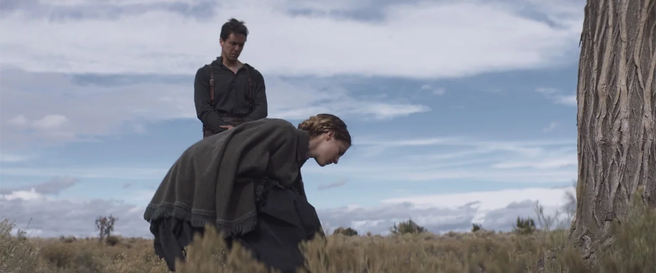 The Wind (2005) movie still: medium — An open field with a large tree under a cloudy blue sky. A man stands behind a woman who is crouched on the…