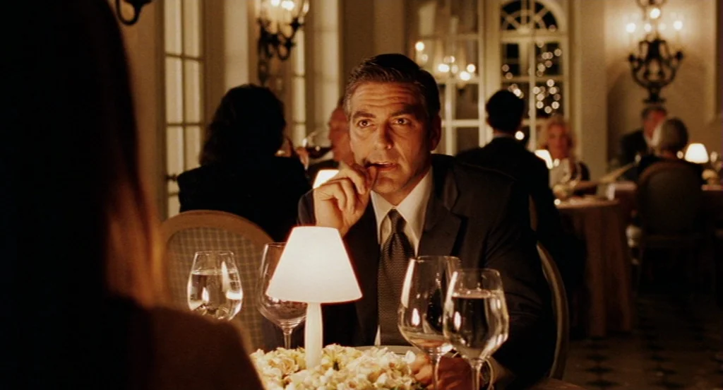 Intolerable Cruelty (2003) movie still: medium — A dimly lit upscale restaurant features tables with small lamps and white flower centerpieces. A…