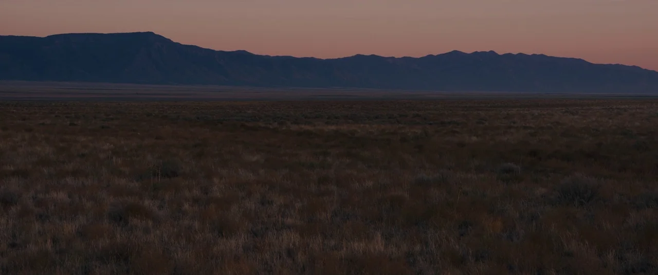 Intrusion (2021) movie still: establishing — A vast, dry desert landscape sits under a faint, warm-toned horizon at dusk. A long, dark mountain…