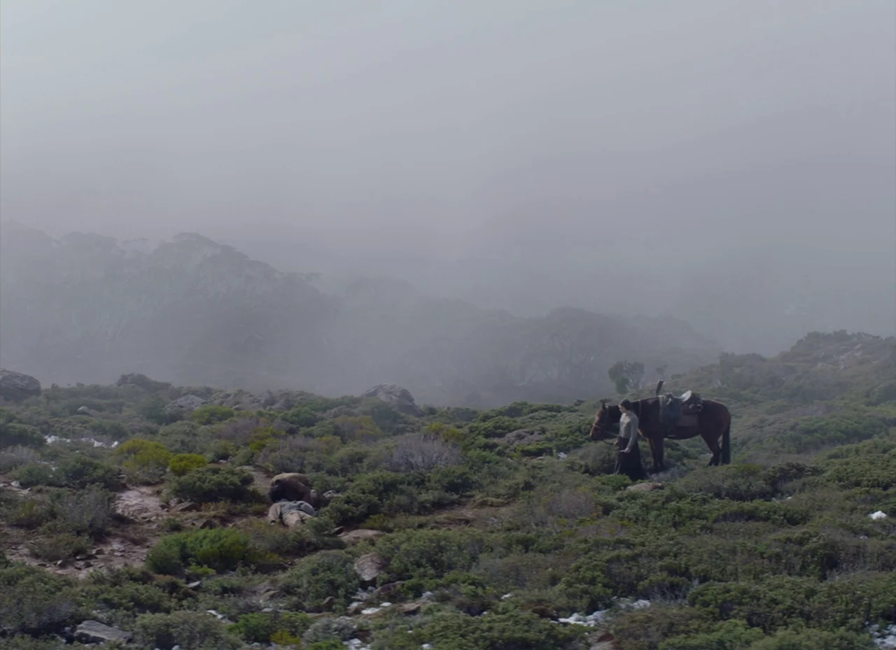 The Nightingale (2018) movie still: wide — A high mountain scrubland is obscured by dense, low-hanging grey fog. A woman stands beside a brown…
