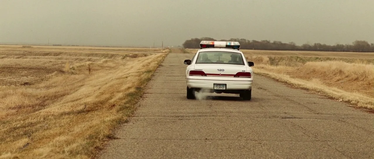 Surveillance (2008) movie still: wide — A flat, desolate rural landscape features dry, golden grass stretching to the horizon. A white police…