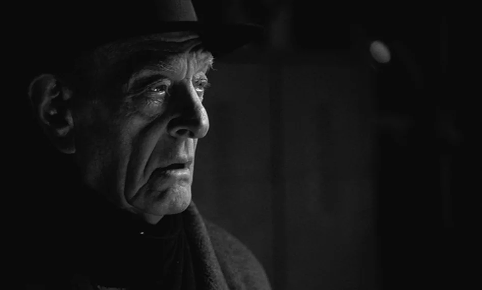 The Man From London (2007) movie still: closeup — A dimly lit room with an elderly man seen in a dark, high-contrast environment. The man looks…