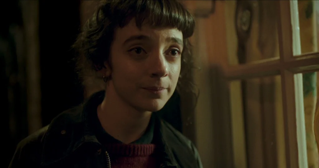 God’s Own Country (2017) movie still: closeup — A dimly lit indoor space with a window frame visible in the foreground. A young woman with short…