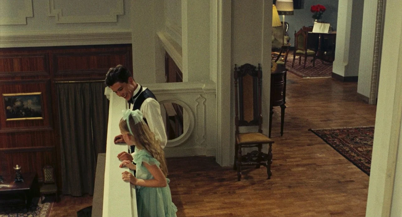 The Damned (1969) movie still: medium — A wooden indoor balcony overlooks a lower level with a high-ceilinged room. A man and a young girl look down…