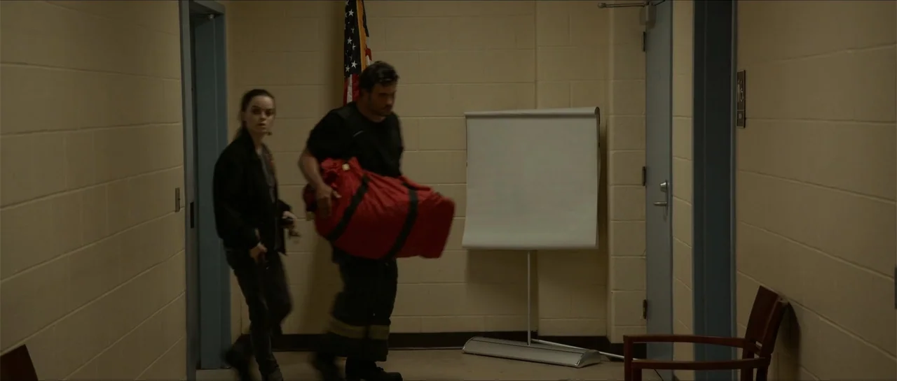 The Vault (2017) movie still: fullbody — A utilitarian office hallway features beige concrete block walls and a standing presentation board. A man…